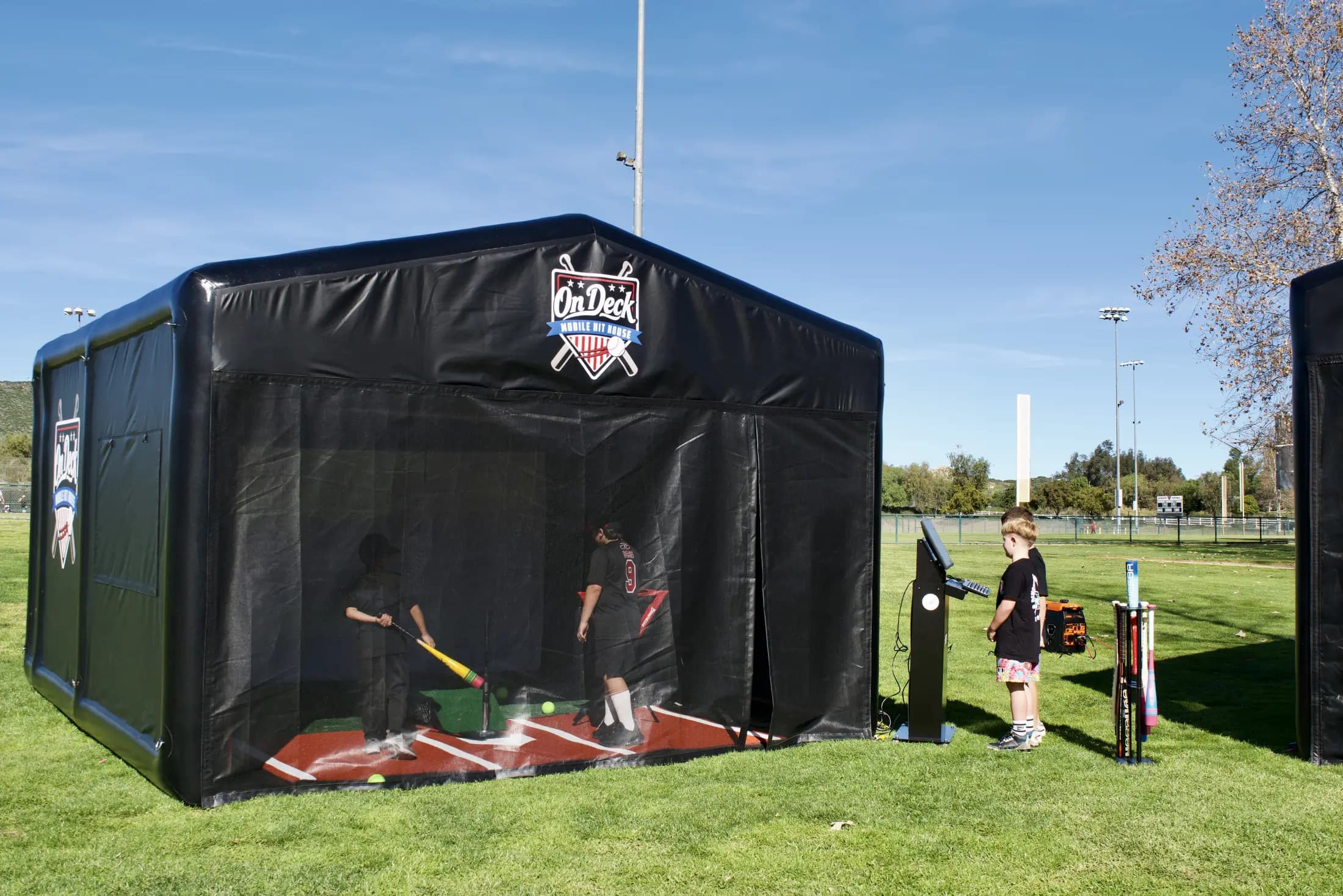 The full On Deck hit house set up on a ball field with a coach and players taking swings